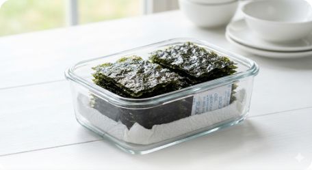 이미지 추천: 유리 밀폐 용기 바닥에 키친타월이 깔려 있고 그 위에 가지런히 잘린 김이 담겨 있는 모습. 옆에는 작은 실리카겔(방습제) 패킷이 놓여 있어 전문적인 느낌을 주는 구도