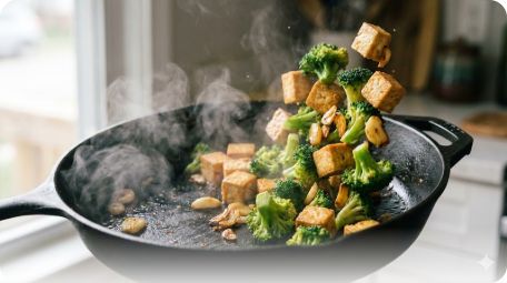 이미지 추천: 김이 나는 프라이팬 안에서 노릇한 두부, 초록색 브로콜리, 황금빛으로 익은 마늘이 조화롭게 섞여 있는 역동적인 요리 사진